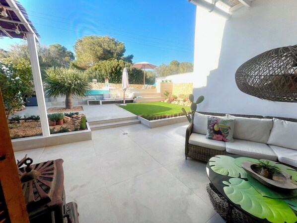 A patio area with a white sofa, a wicker chair, and a decorative plant on a table. There is a small garden with a lawn and a stone wall in the background.