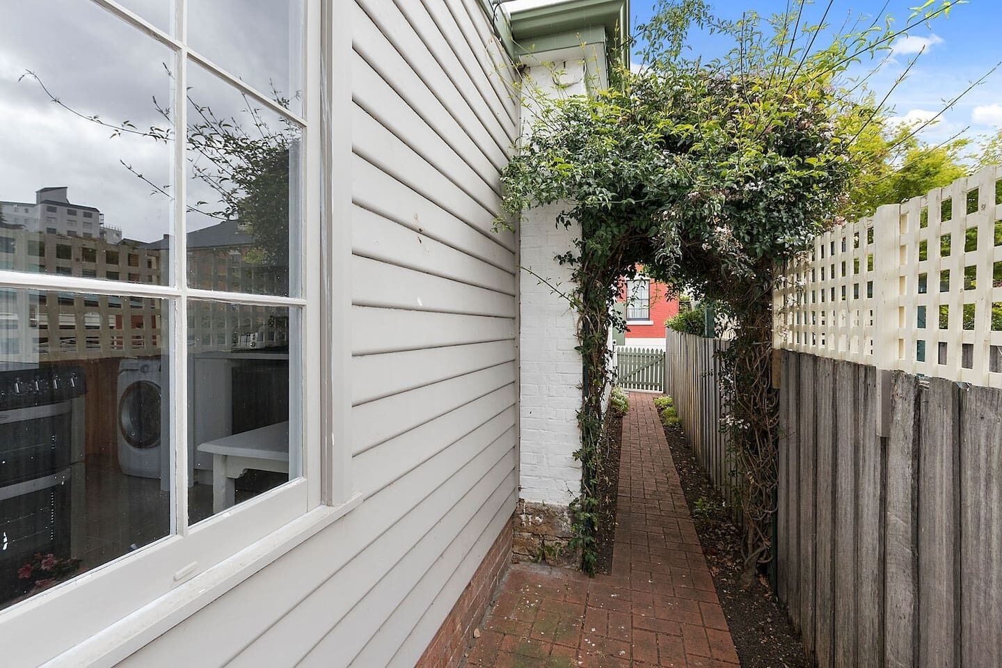 A narrow brick pathway leading to a wooden fence, with a white building on one side and a red building in the background.