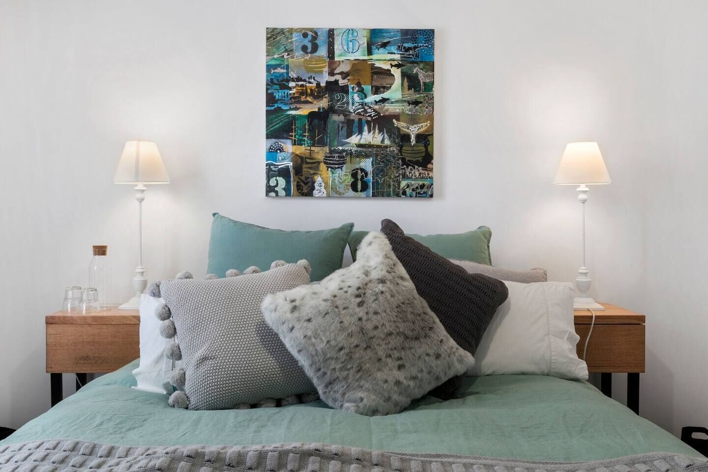 A neatly made bed with a green bedspread, multiple pillows, and two bedside tables with lamps. A colorful abstract artwork is displayed above the bed.