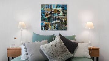 A neatly made bed with a green bedspread, multiple pillows, and two bedside tables with lamps. A colorful abstract artwork is displayed above the bed.