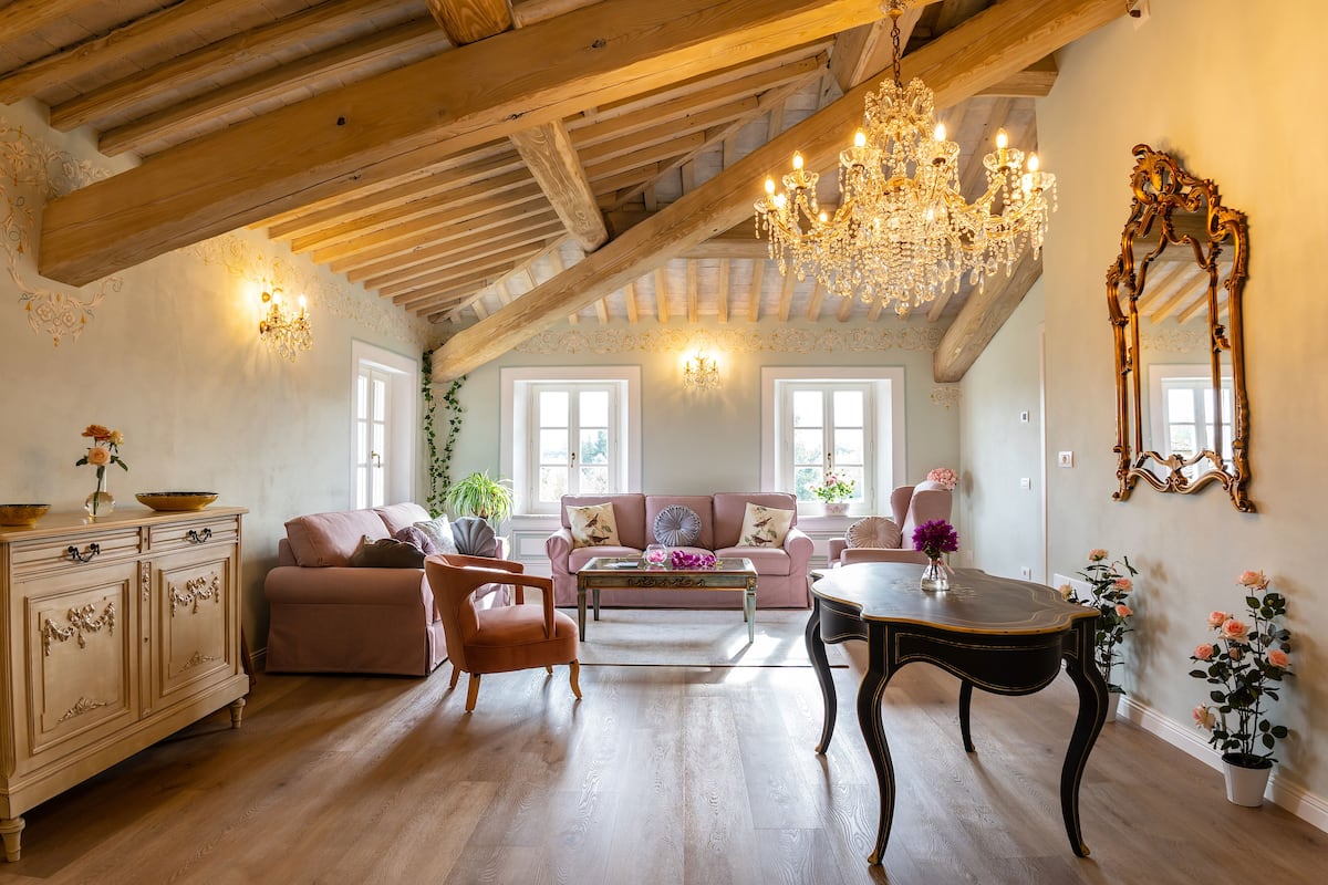 A spacious living room with a high ceiling, wooden beams, a chandelier, a pink sofa, a coffee table, and a dining table.