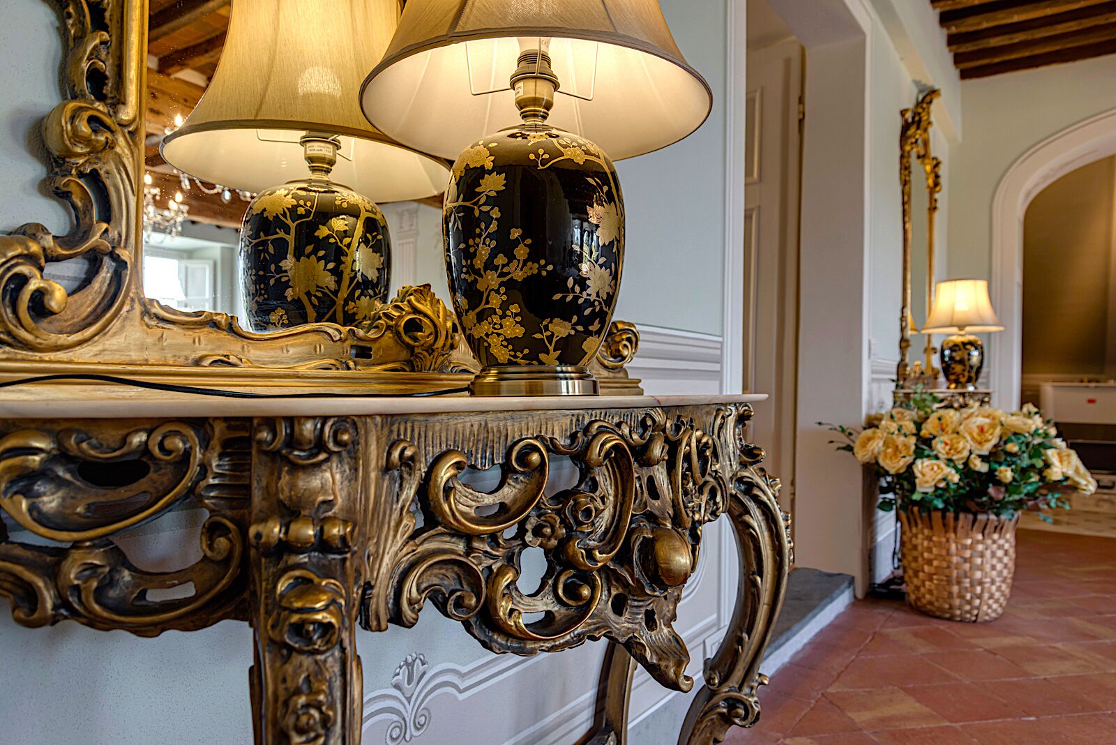 An ornate gold and black console table with a lamp and a vase, set against a white wall with a mirror.
