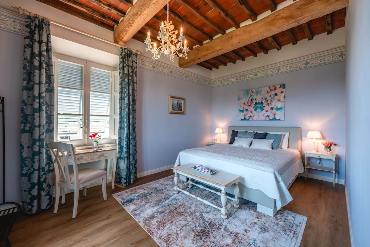 A bedroom with a wooden ceiling, a bed with white bedding, a desk with a chair, and a window with curtains.