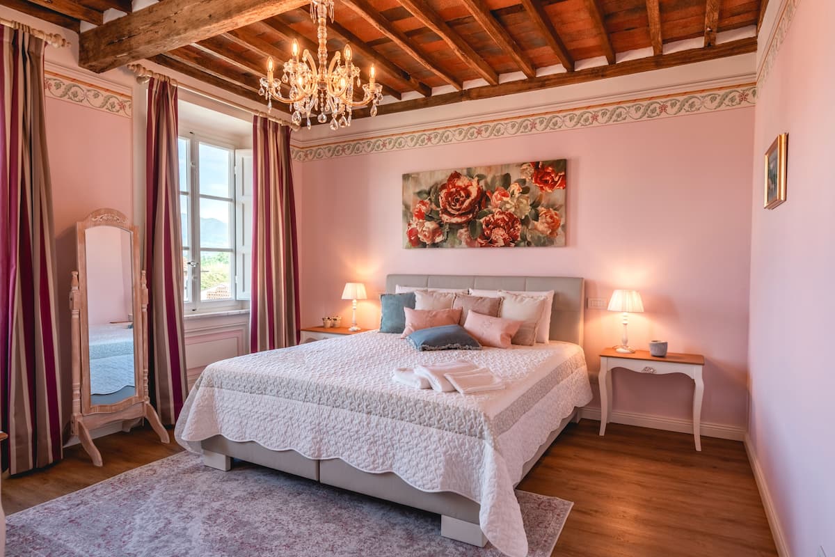 A bedroom with a large bed, two bedside tables, a chandelier, and a floral painting on the wall.