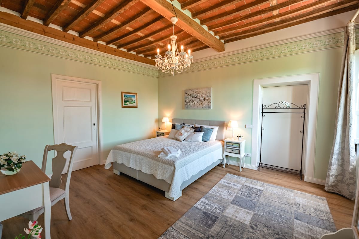 A bedroom with a wooden ceiling, a chandelier, a bed with white bedding, a bedside table, a chair, and a door.