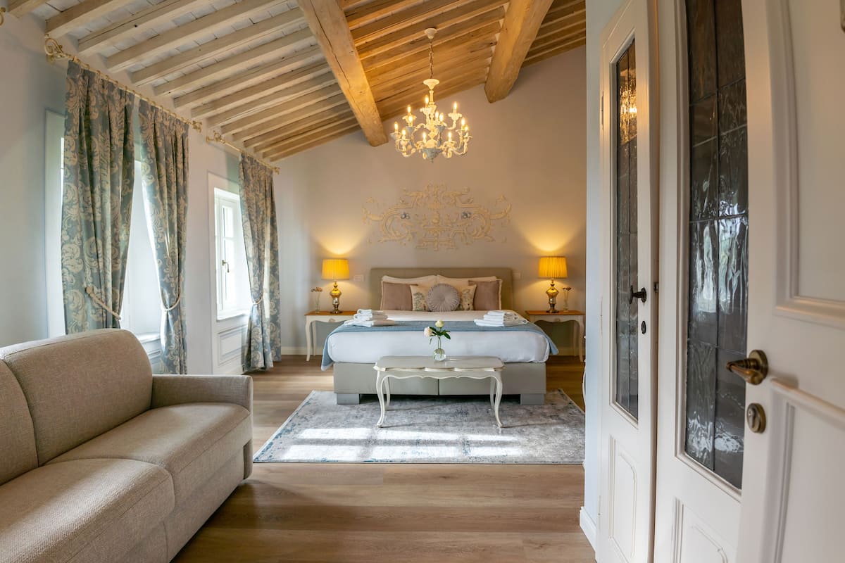 A bedroom with a bed, bedside lamps, a chandelier, a sofa, and a door.