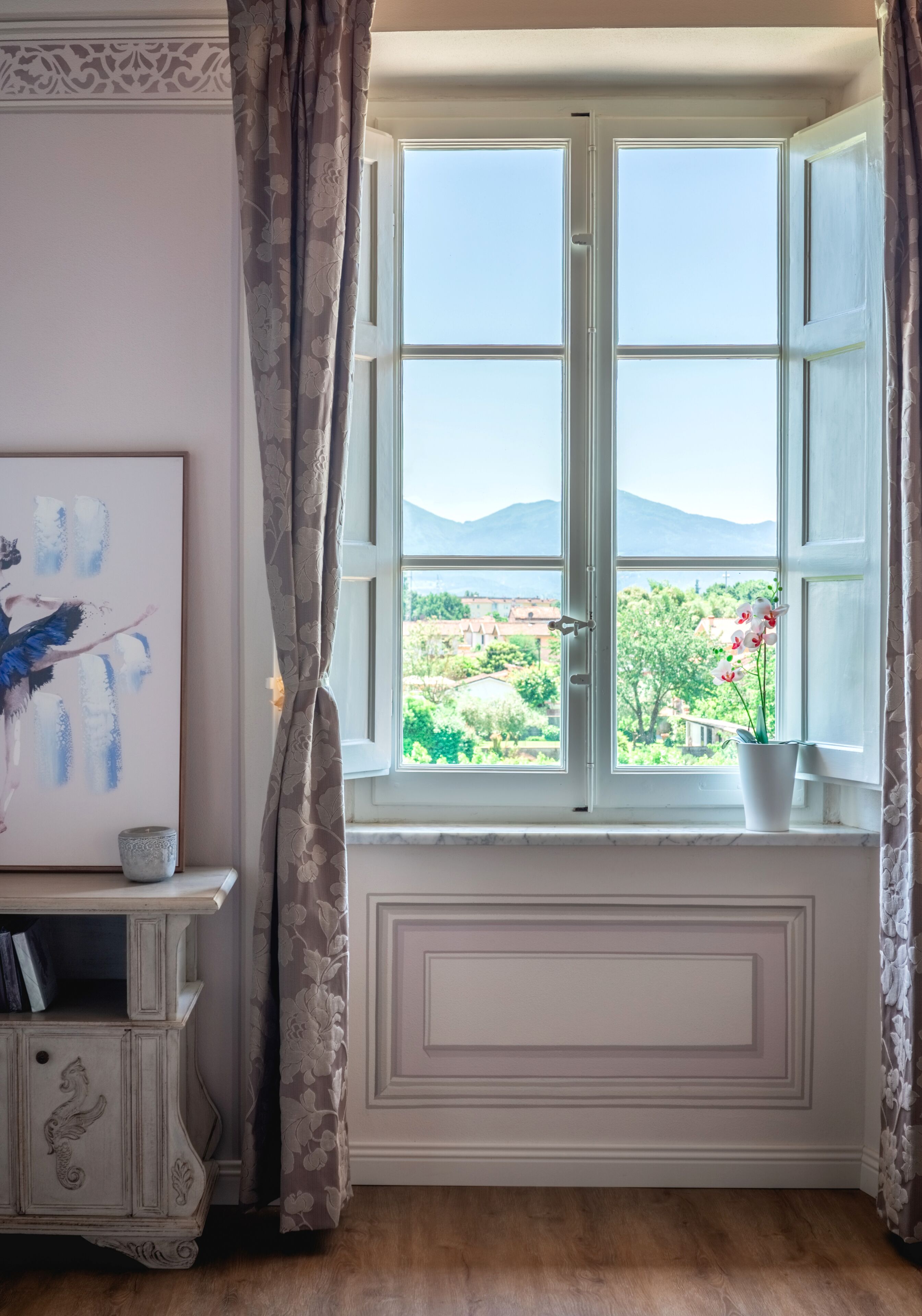 A room with a large window, a painting on the wall, and a small table with a vase of flowers.