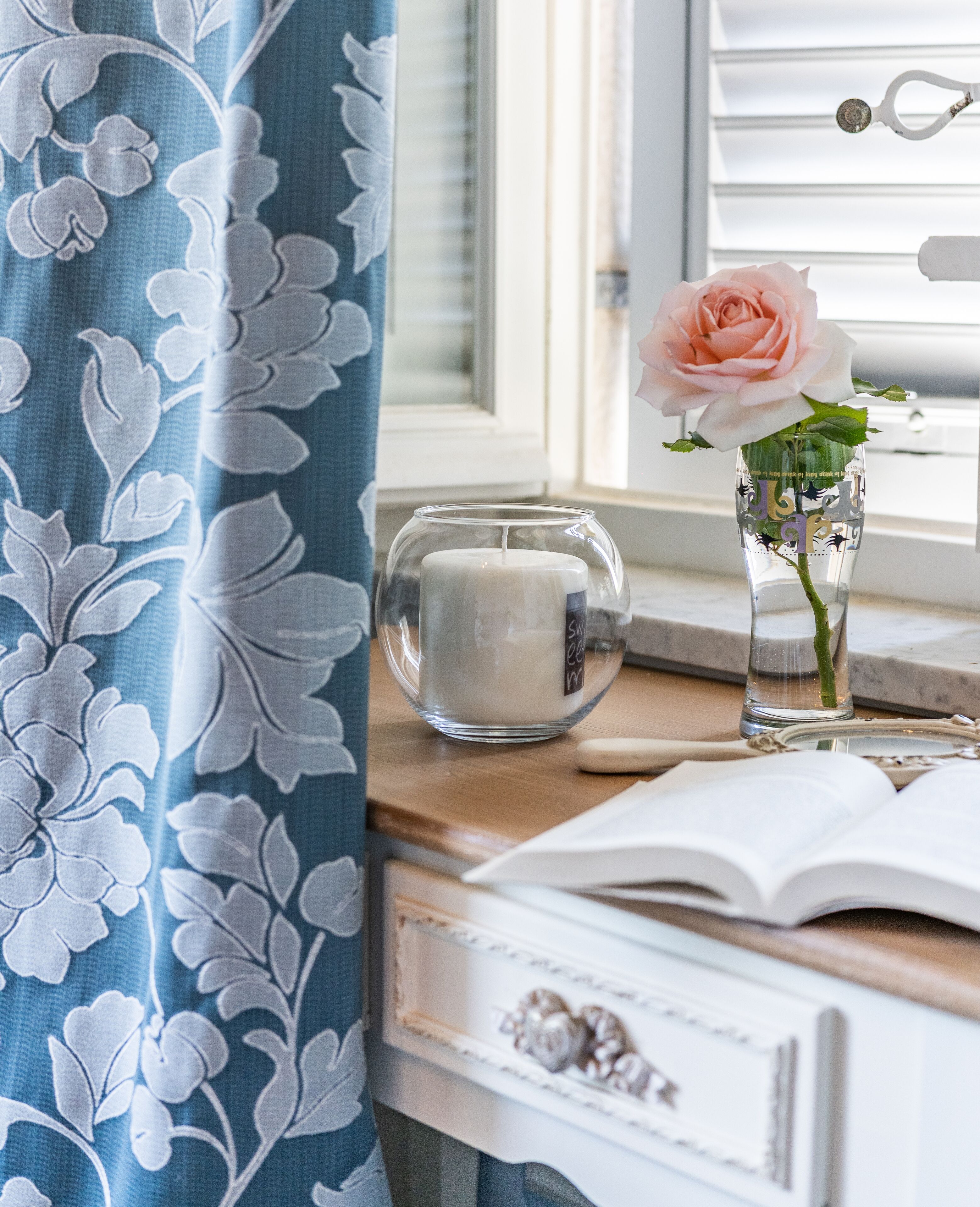 A room with a window, a candle, a vase with a rose, and an open book on a wooden table.