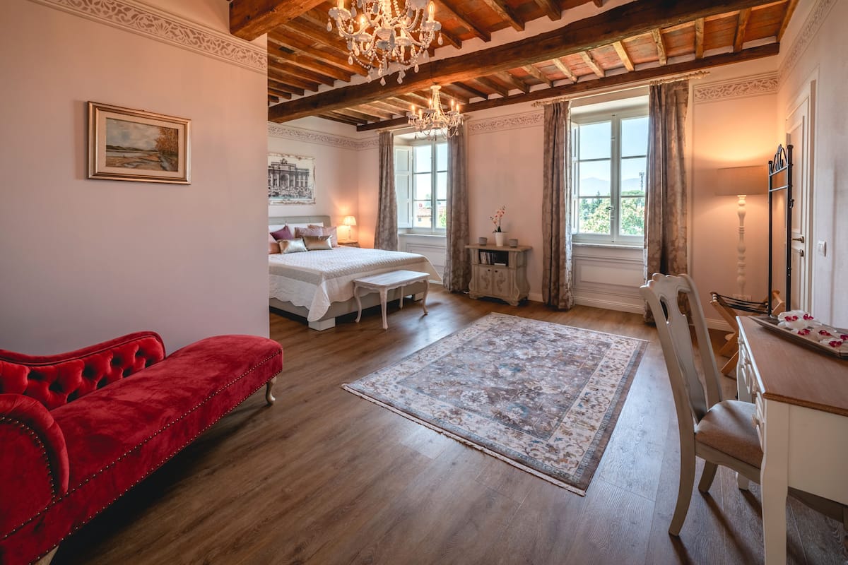 A spacious room with a wooden ceiling, a large bed, a red sofa, a dining table, and a view of the outdoors.