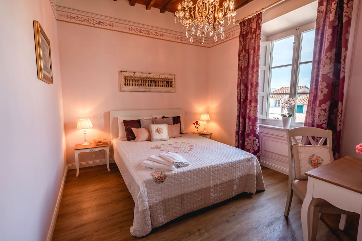 A bedroom with a bed, bedside lamps, a chandelier, a window with curtains, and a small table with a chair.