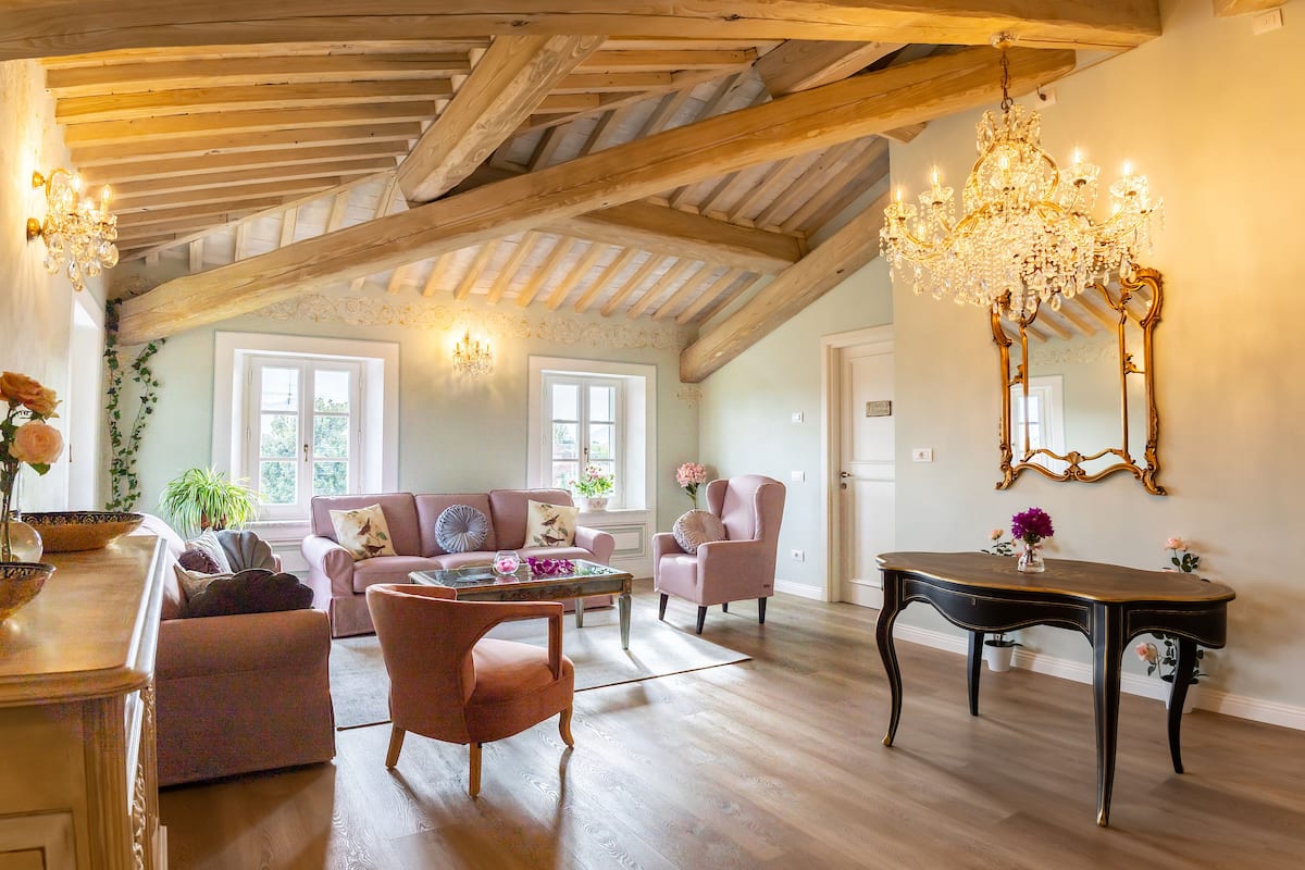 A spacious living room with a high ceiling, wooden beams, a chandelier, a sofa, a chair, a coffee table, and a mirror.