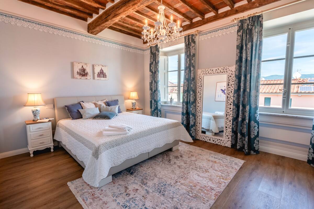 A bedroom with a large bed, two bedside tables with lamps, a chandelier, a large window with curtains, and a view of buildings outside.
