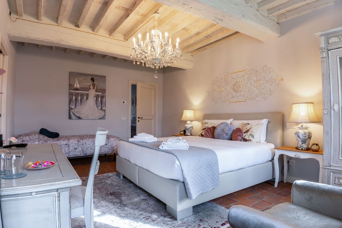 A bedroom with a large bed, two bedside tables with lamps, a chandelier, a painting on the wall, and a small table with a vase.