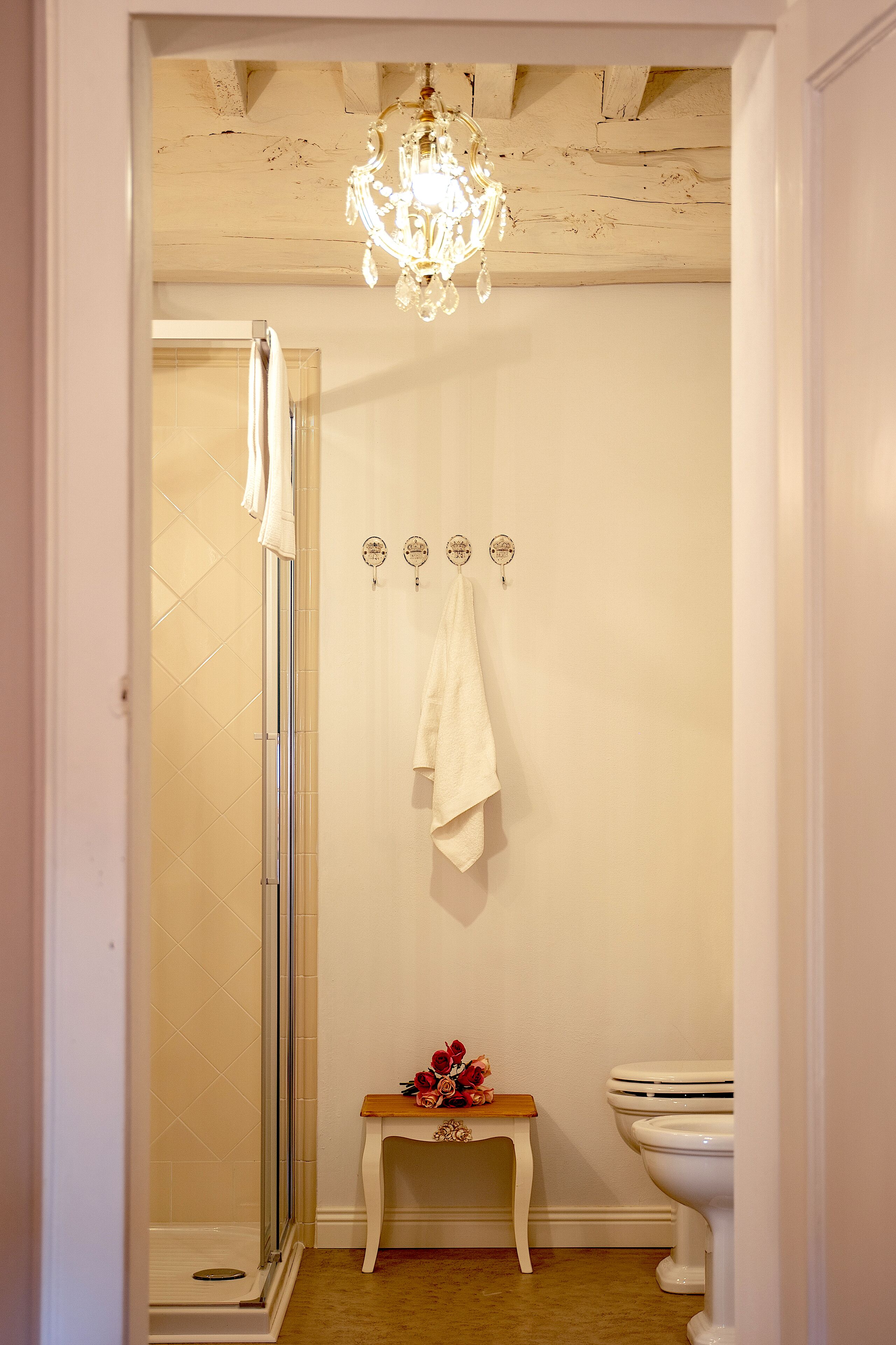 Een badkamer met een glazen douchedeur, een wit toilet, een bankje met bloemen en een kroonluchter.
