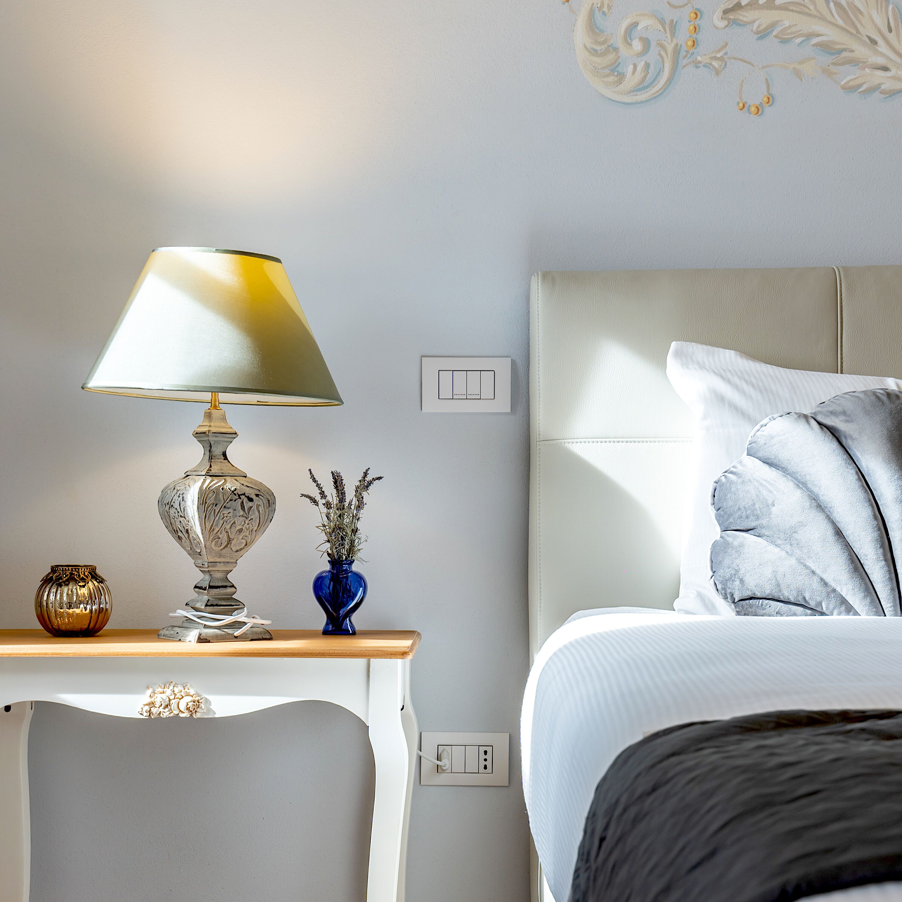 A neatly arranged bedroom with a bed, bedside table, lamp, vase with flowers, and a wall switch.