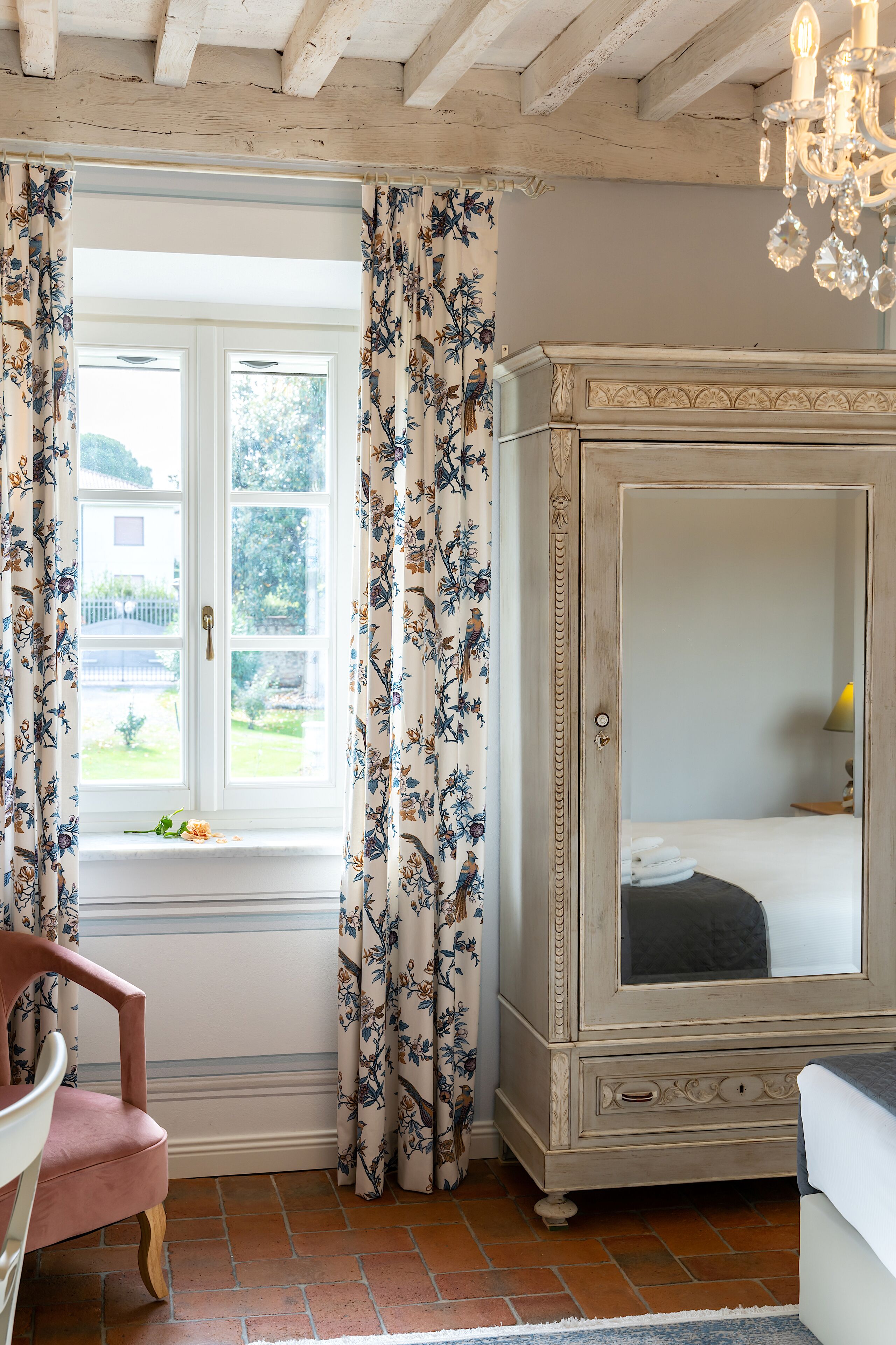 A bedroom with a large window, floral curtains, a wooden cabinet, and a chandelier.