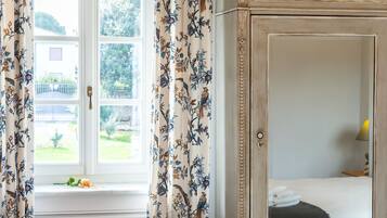 A bedroom with a large window, floral curtains, a wooden cabinet, and a chandelier.