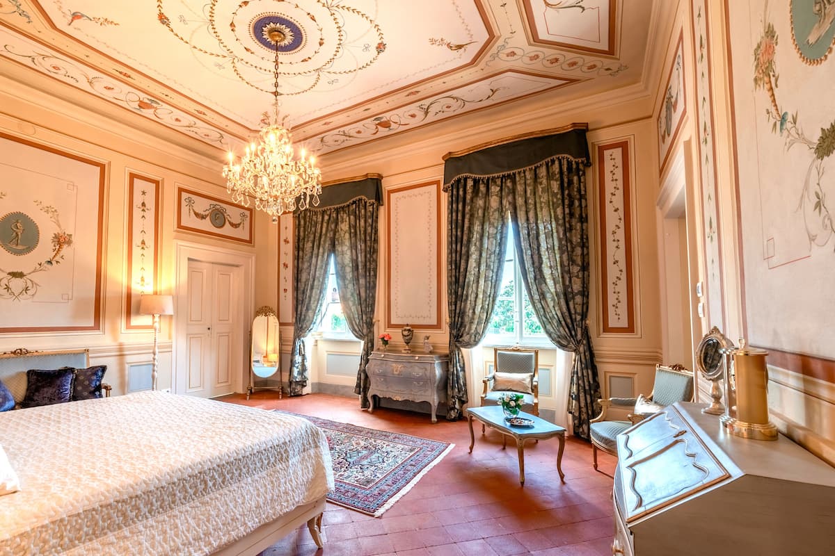 A luxurious room with a large bed, a chandelier, a fireplace, and ornate wall decorations.