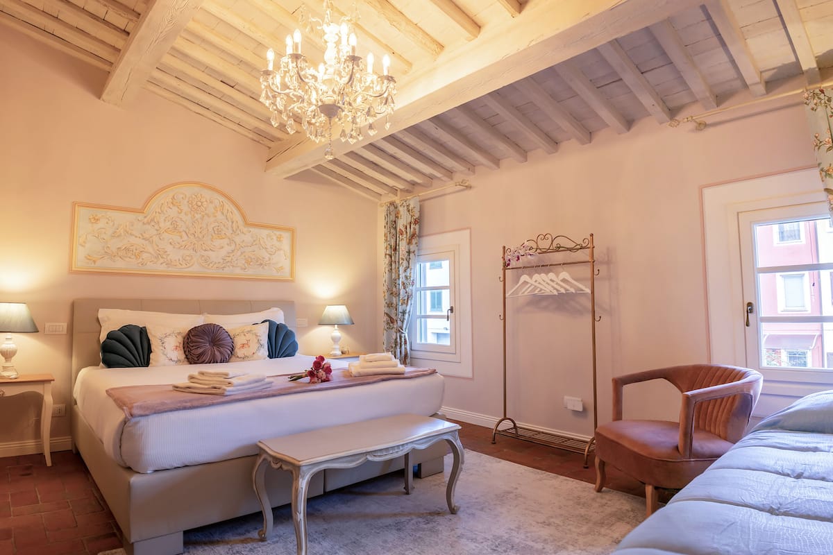 A bedroom with a large bed, a bench, a chair, a chandelier, and a wardrobe.