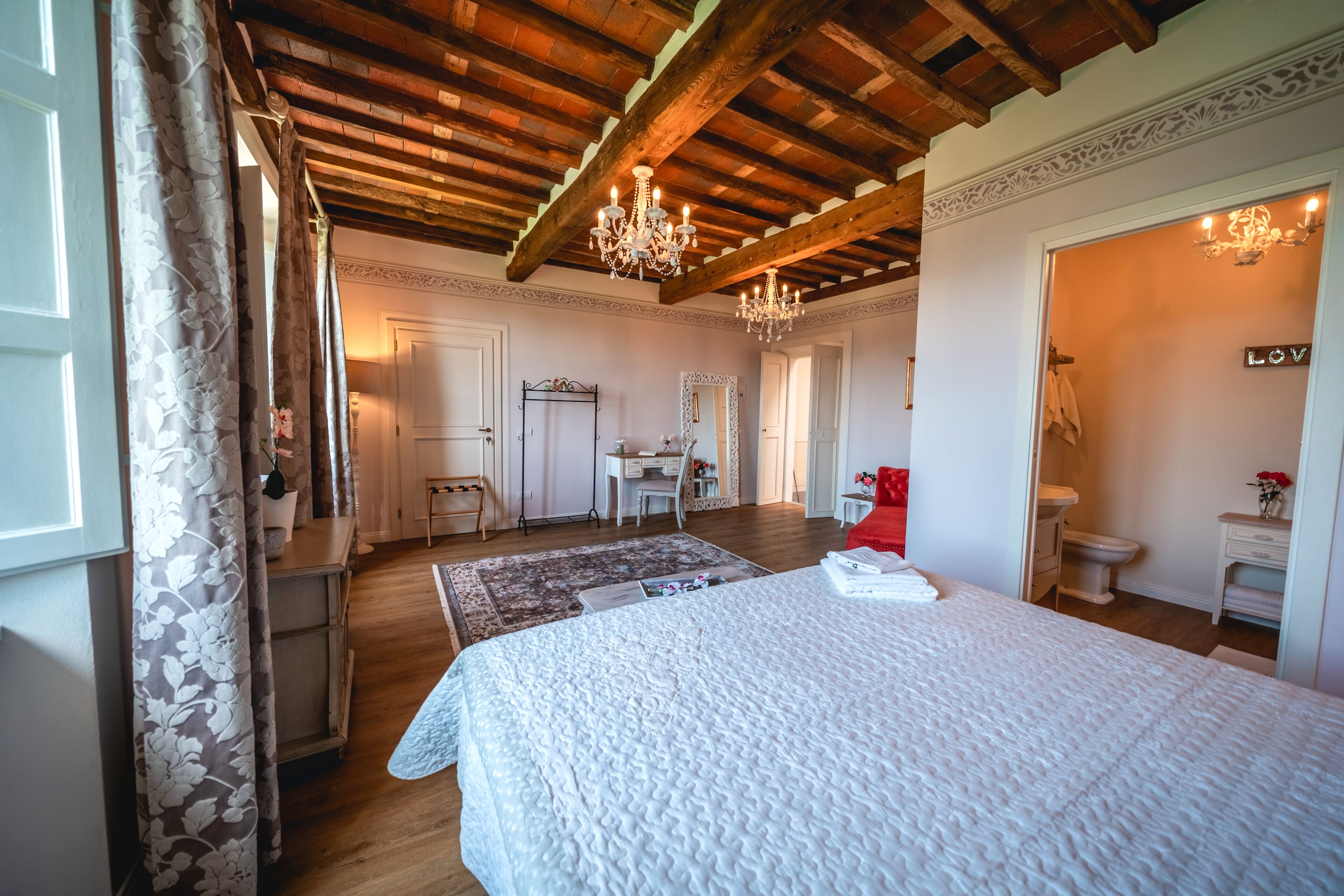 A bedroom with a large bed, wooden ceiling beams, a desk, and a bathroom with a toilet and sink.