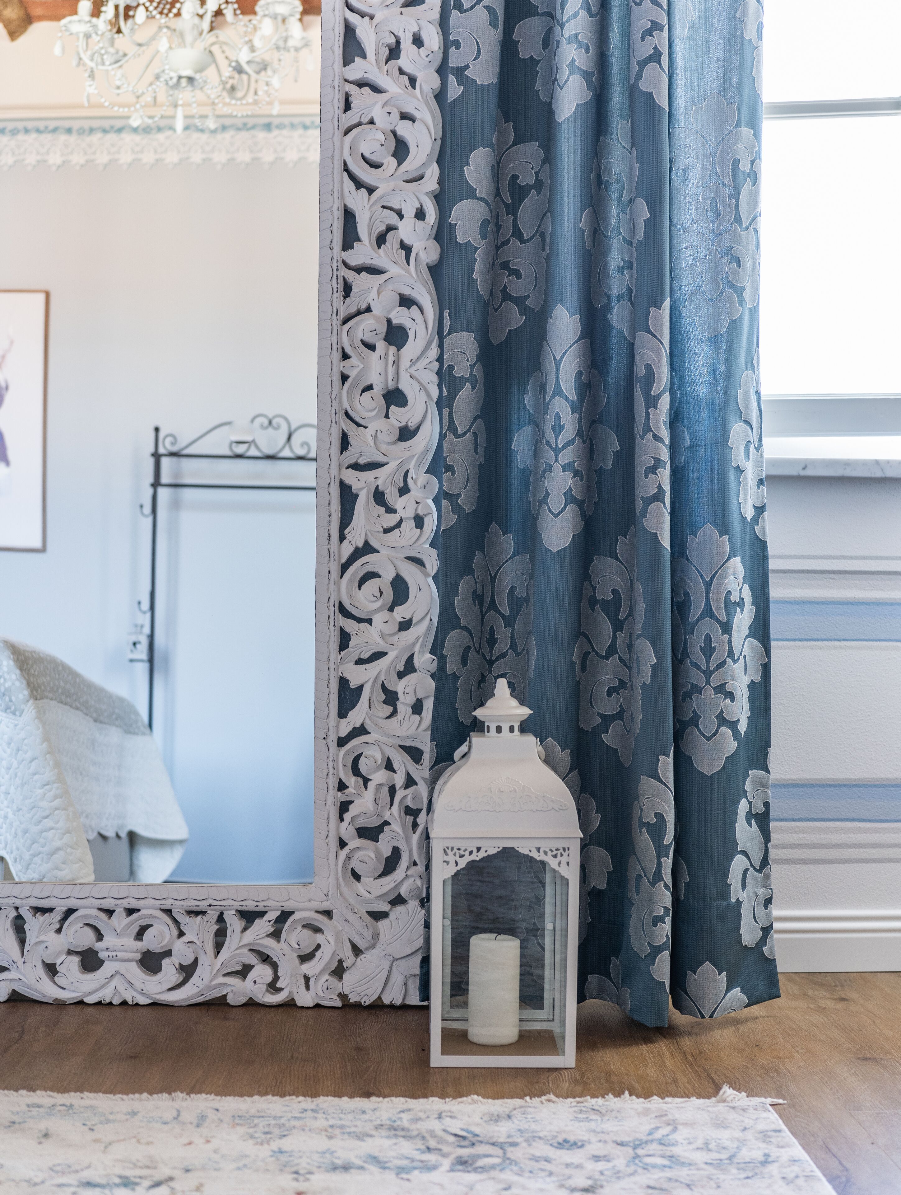 Una habitación con cortinas blancas ornamentadas, un farol de marco blanco y una alfombra estampada.