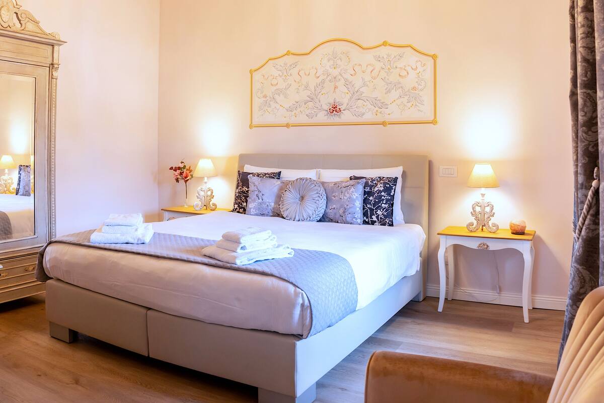 A well-lit bedroom with a large bed, bedside lamps, a chandelier, and a decorative wall piece.
