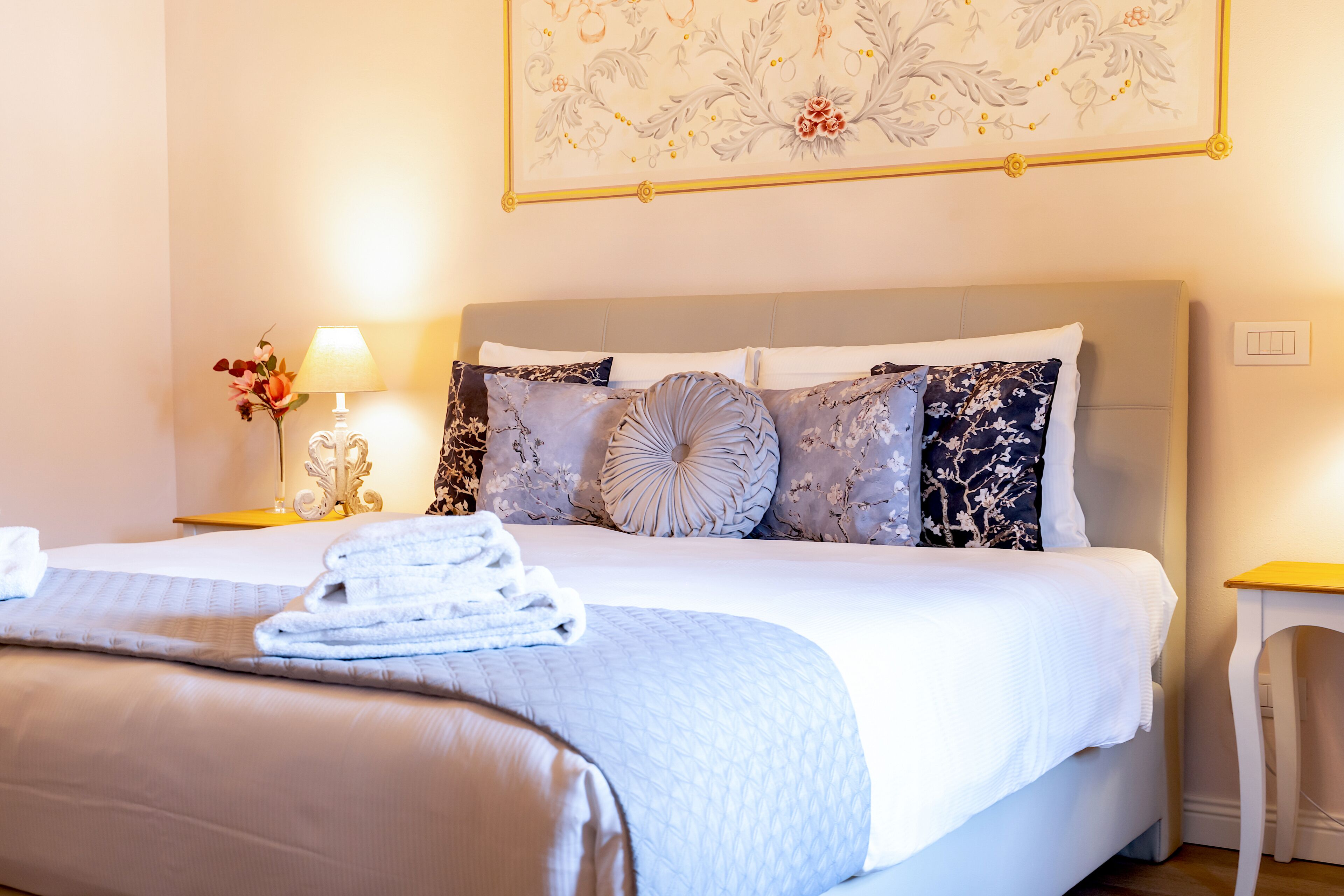 Een keurig opgemaakt bed met een grijze hoofdbord, een bloemrijk kunstwerk erboven en een nachtkastje met een lamp en een bloemenvaas.