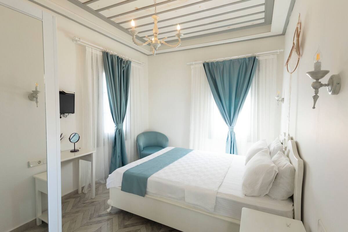 Un dormitorio con cama, mesita de noche, silla, espejo y ventana con cortinas.