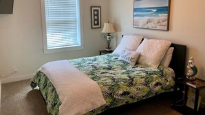 Een slaapkamer met een bed, een plafondventilator, een raam met zonwering, een nachtkastje met een lamp en een schilderij aan de muur.