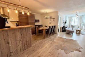 A modern kitchen with wooden cabinets, a dining area with a wooden table and chairs, and a living space with a sofa and a coffee table.