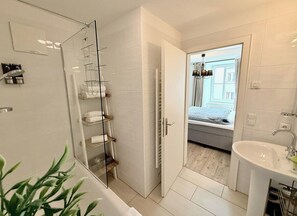 Ein modernes Badezimmer mit Duschkabine aus Glas, einem weißen Waschbecken und Blick auf ein Schlafzimmer durch eine geöffnete Tür.