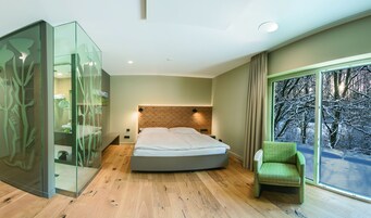 Un dormitorio moderno con una cama grande, un separador de vidrio, una silla verde y vistas a un bosque nevado a través de un ventanal.