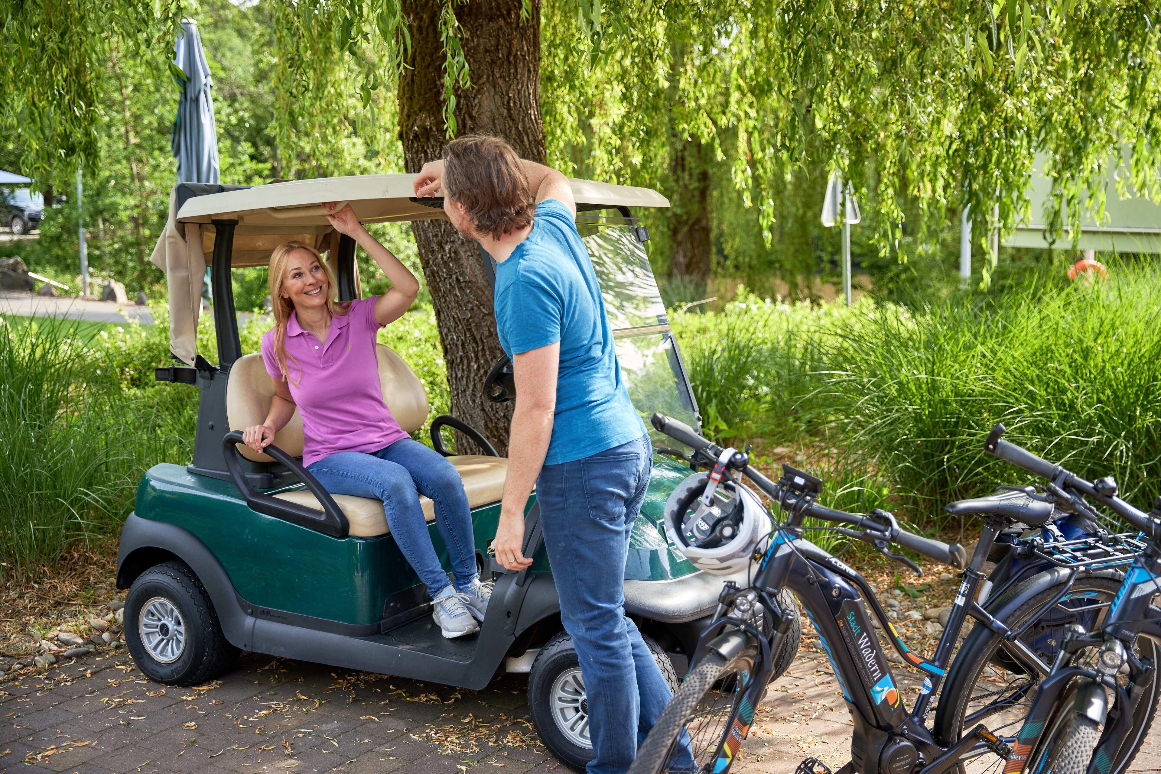 Une voiturette de golf, avec une personne debout à côté, une autre assise à l’intérieur, et un vélo garé à proximité.