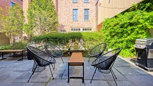 Une terrasse extérieure avec une table en bois et quatre chaises en fil de fer noir, entourée de carreaux de pierre et de verdure.