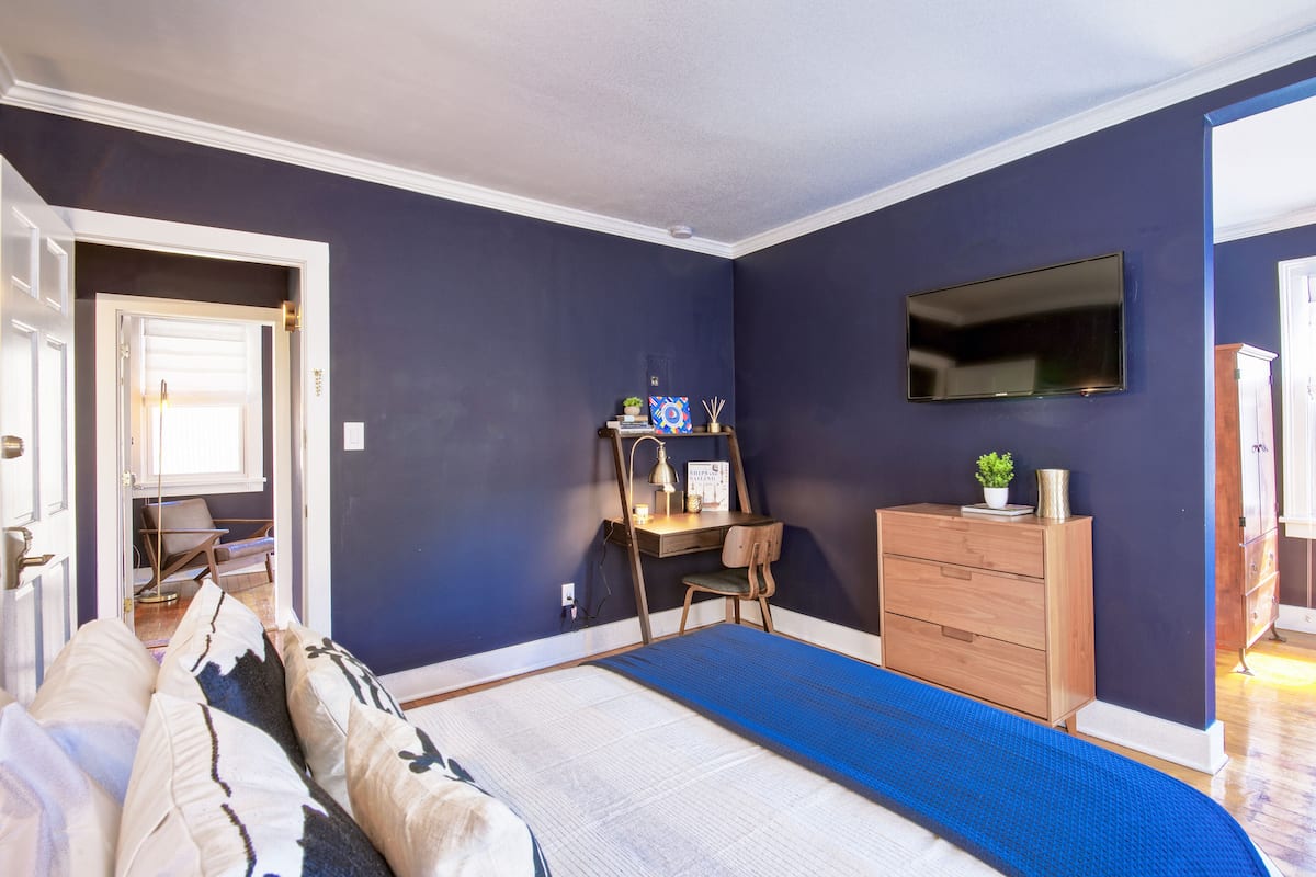 A bedroom with a bed, a desk, a dresser, and a TV on the wall.