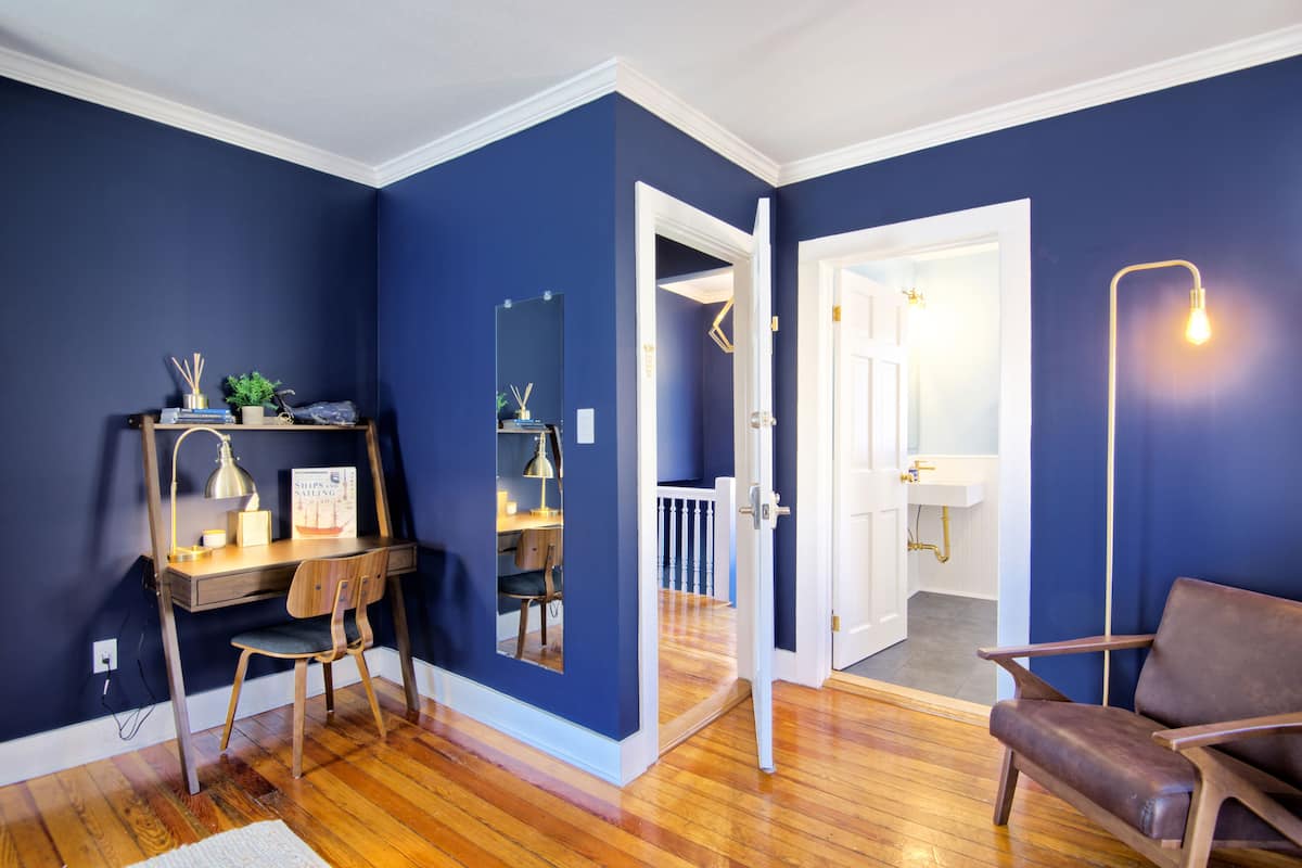 A room with a desk, chair, and a brown leather chair. There is a white door leading to a bathroom with a sink and a mirror.
