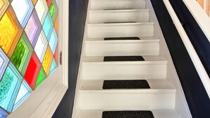 Un escalier avec des marches blanches et des contremarches noires antidérapantes, menant à un palier avec un mur sombre et une fenêtre ornée de vitraux colorés.