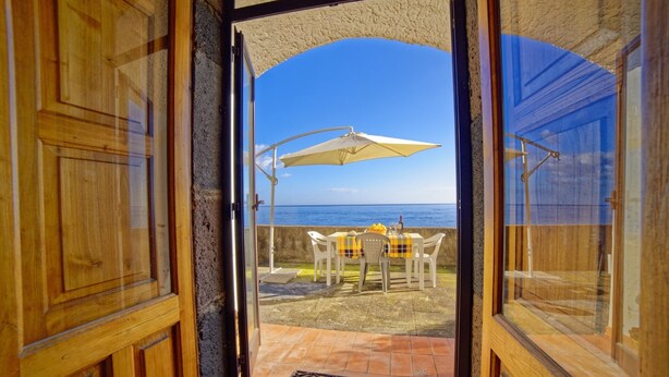 Une porte ouverte donnant sur une terrasse avec une table et des chaises, un parasol blanc et une vue sur la mer.