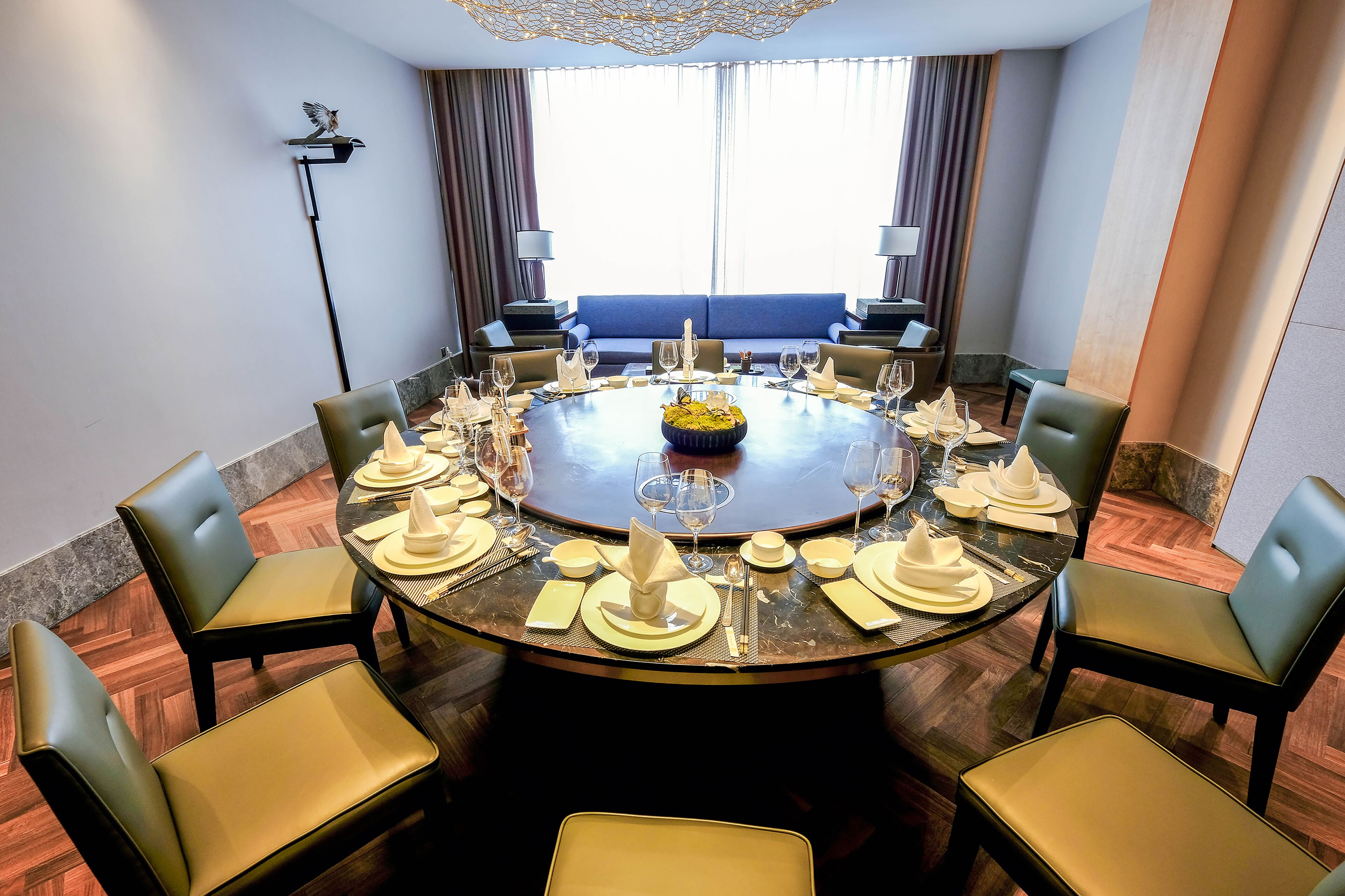 A round dining table set for eight with chairs, a central floral arrangement, and a view of a blue sofa through the window.
