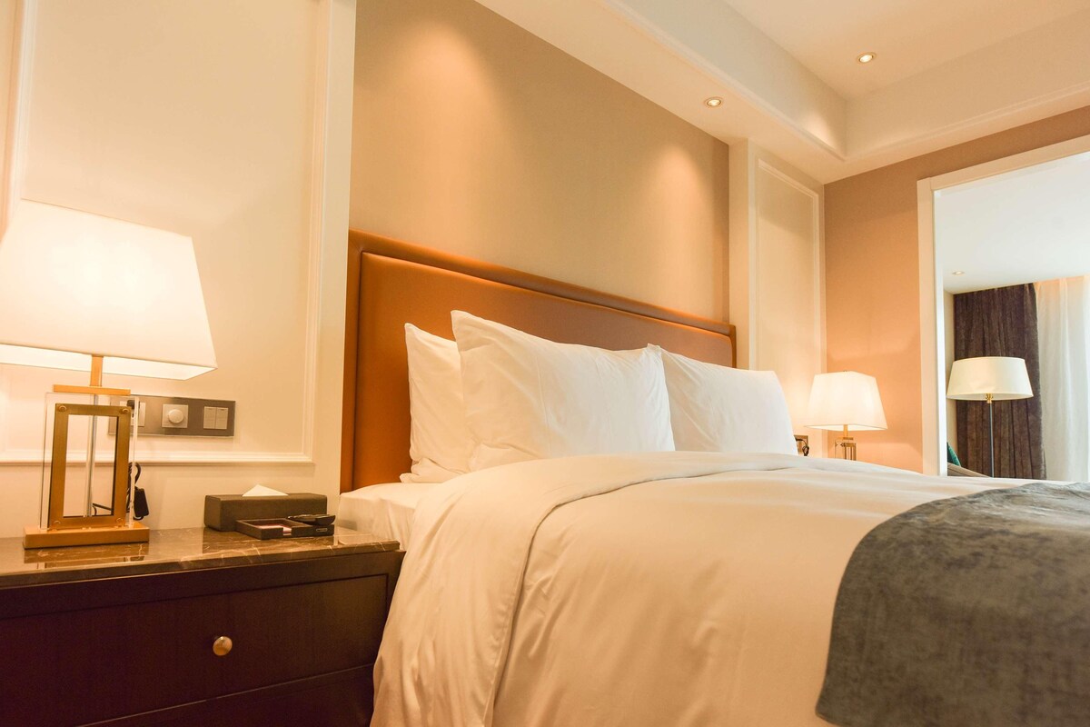 A hotel room with a bed, nightstand, lamp, and a view of the outside.