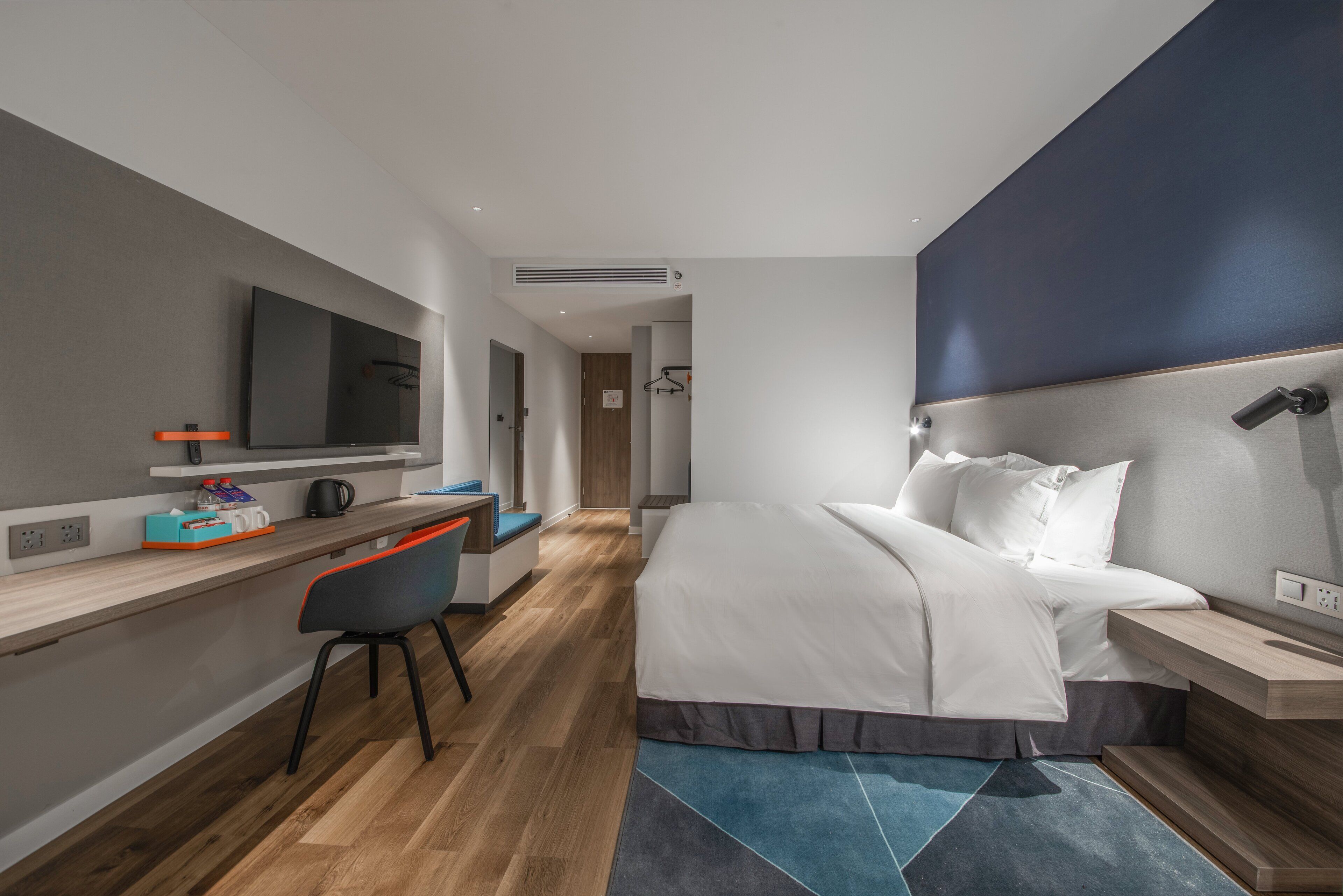A modern hotel room with a bed, desk, chair, and a large flat-screen TV.