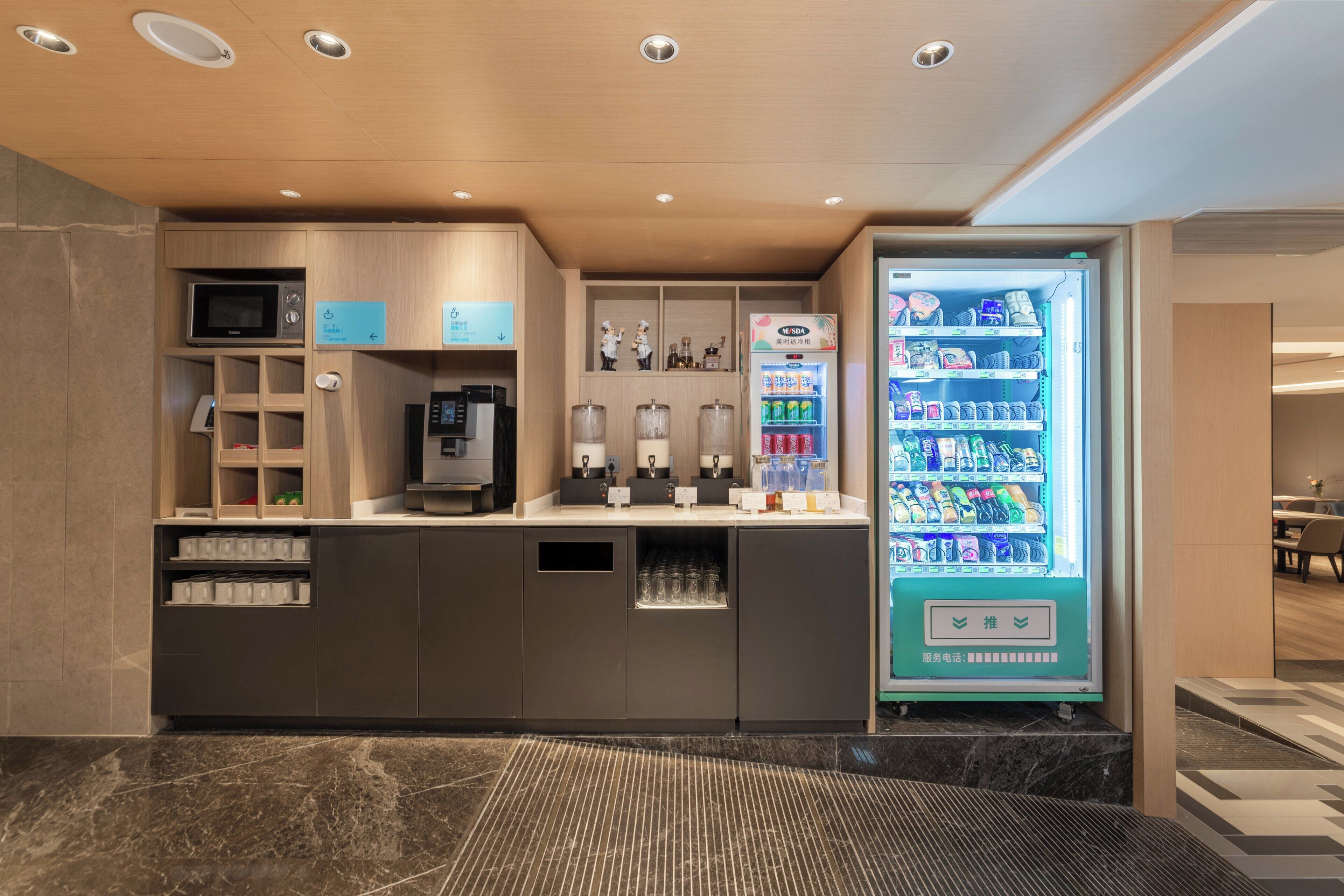 Un área de recepción de hotel moderna con una máquina expendedora de bebidas, una cafetera y un microondas.