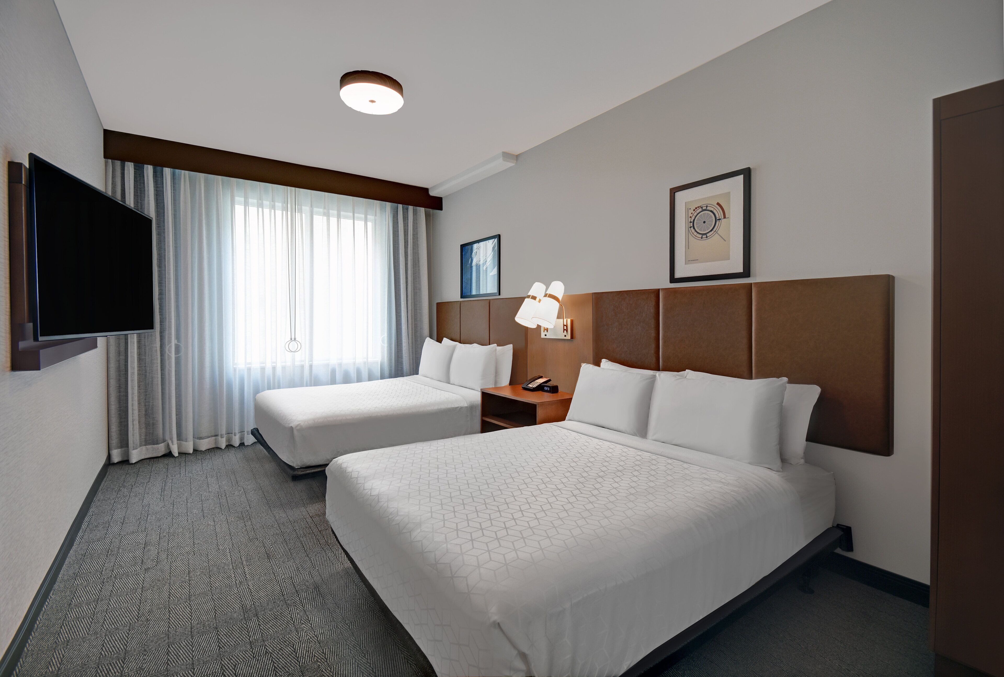 Une chambre d’hôtel avec deux lits, un téléviseur à écran plat, une lampe de chevet, une horloge et une fenêtre avec des rideaux.