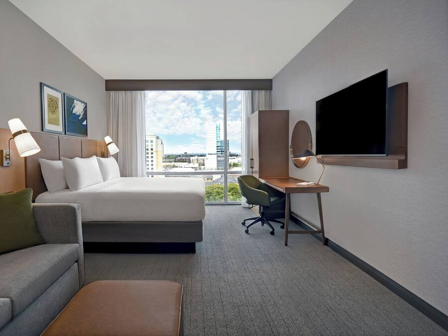 A hotel room with a bed, a desk, a television, and a view of the cityscape.