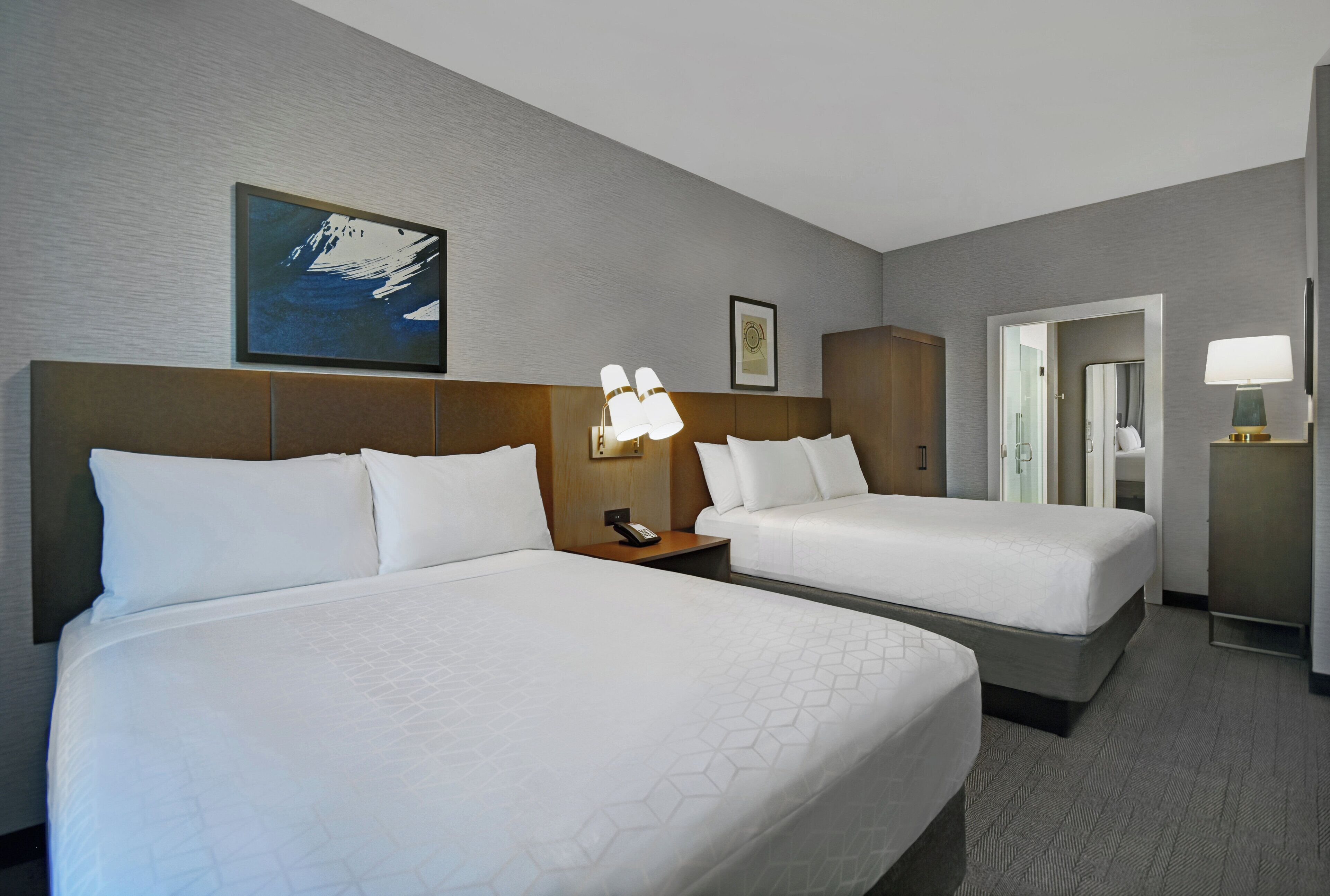 Ein Hotelzimmer mit zwei Betten, einem Nachttisch mit Lampe, einem gerahmten Bild an der Wand und einer Tür zu einem Badezimmer.