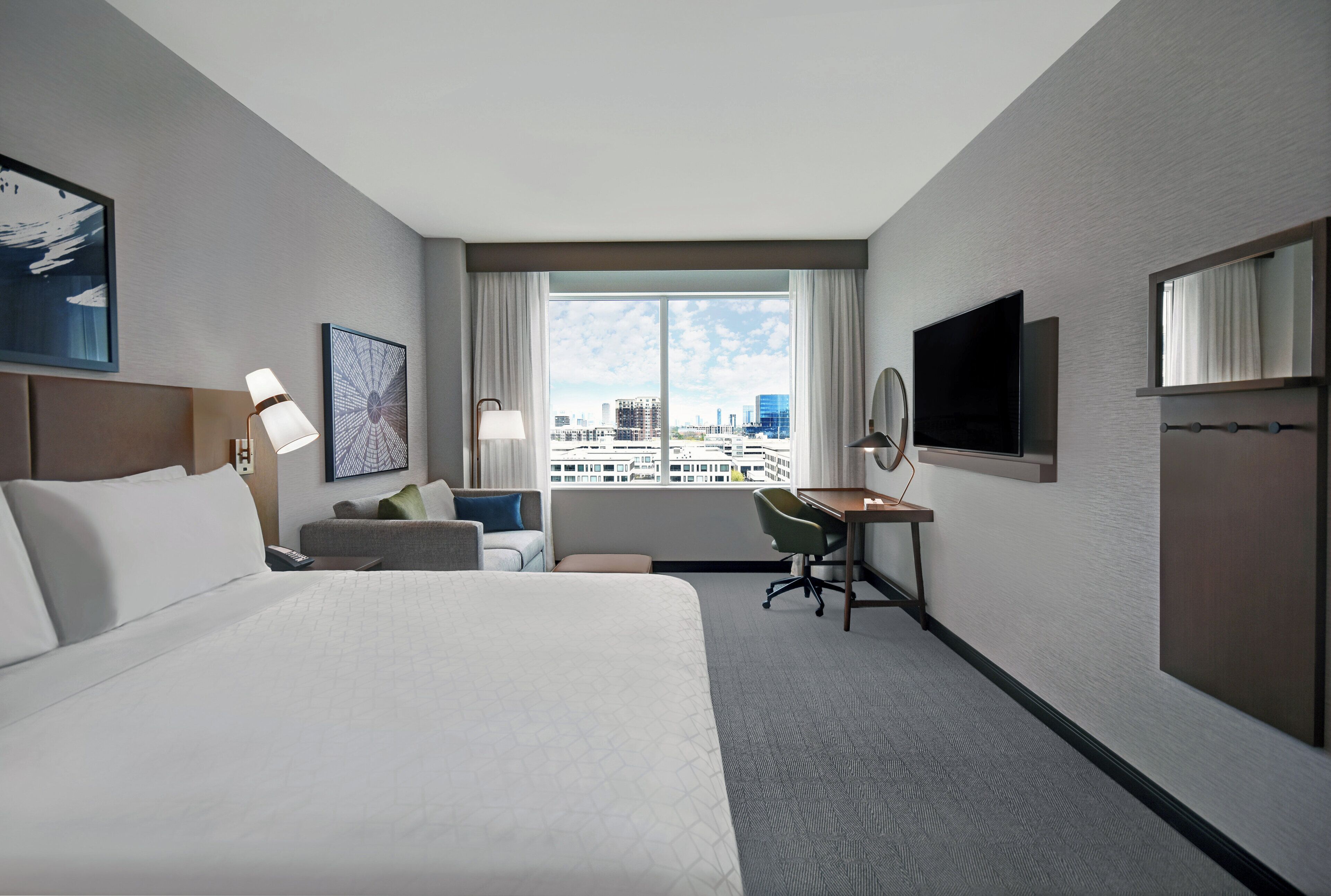 A hotel room with a large bed, a desk, a chair, a TV, and a window with a city view.