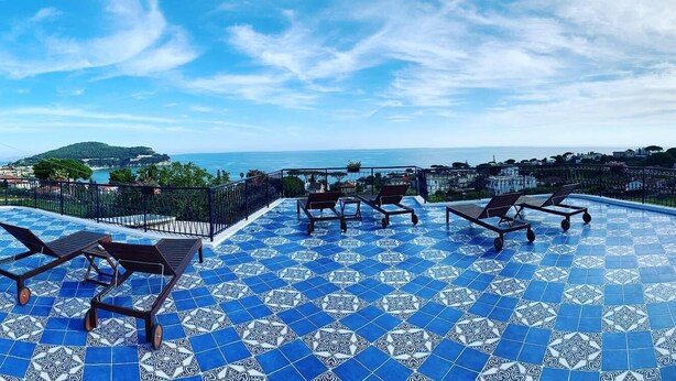 A rooftop pool with blue patterned tiles, sun loungers, and a view of the sea and distant hills. - Gli Archi sul Mare (Gaeta)