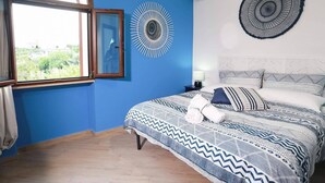 A bedroom with a large bed, a window with curtains, a wall-mounted air conditioner, and a decorative wall piece. - Gli Archi sul Mare (Gaeta)
