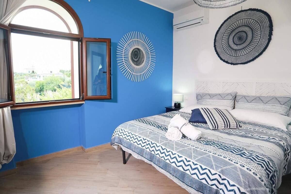 Un dormitorio con una cama grande, una ventana con cortinas, un aire acondicionado de pared y un adorno decorativo en la pared.