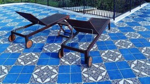 A terrace with blue patterned tiles, two wooden lounge chairs, and a view of the sea and a distant town. - Gli Archi sul Mare (Gaeta)